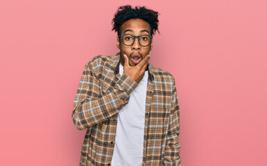 Young african american man with beard wearing casual clothes and glasses looking fascinated with disbelief, surprise and amazed expression with hands on chin