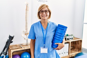 Middle age blonde woman wearing physiotherapist uniform holding foam roller at clinic