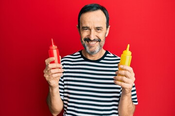 Middle age hispanic man holding ketchup and mustard bottle smiling with a happy and cool smile on...