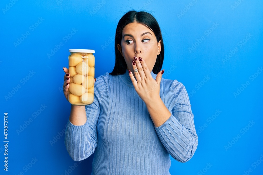 Poster Beautiful brunette woman holding bottle with boiled potatoes covering mouth with hand, shocked and afraid for mistake. surprised expression