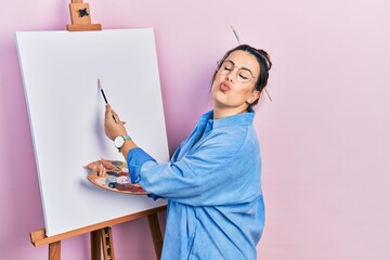 Young hispanic woman standing drawing with palette by painter easel stand looking at the camera...