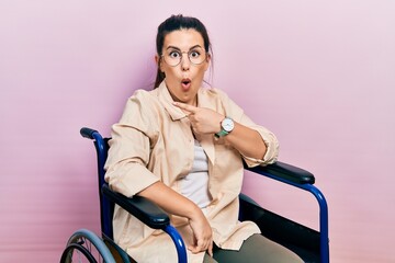 Obraz na płótnie Canvas Young hispanic woman sitting on wheelchair surprised pointing with finger to the side, open mouth amazed expression.