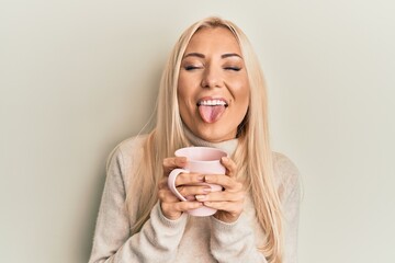 Young blonde woman drinking a cup of coffee sticking tongue out happy with funny expression. © Krakenimages.com