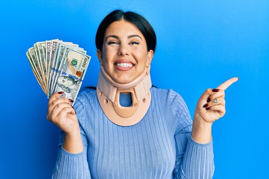 Beautiful brunette woman wearing cervical collar holding insurance money smiling happy pointing with hand and finger to the side