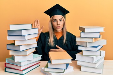 Young caucasian woman wearing graduation ceremony robe sitting on the table swearing with hand on chest and open palm, making a loyalty promise oath
