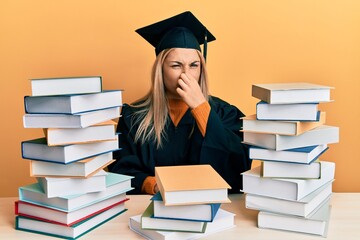 Young caucasian woman wearing graduation ceremony robe sitting on the table smelling something stinky and disgusting, intolerable smell, holding breath with fingers on nose. bad smell - obrazy, fototapety, plakaty