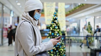 Young Caucasian Woman in Fashionable Clothes and Surgical Hygiene Mask Waiting in Shopping Mall Swiping Her Smartphone Looking for Best Offers on Christmas Season Sale Day	