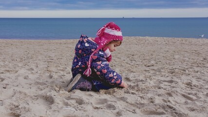 Young Curious Caucasian Girl in Warm Winter Jumpsuit Searching for Seashells and Amber Rock in Beach Sand on Cold Autumn Winter Day	