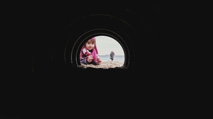 Curious Caucasian Girl in Warm Clothes Looking and Crawling into Dark Narrow Tunnel on Beach	
