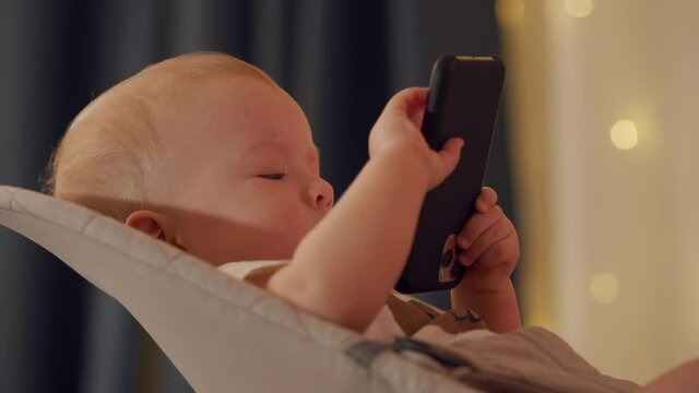 Baby boy sitting on rocking chair for babies playing with smartphone, cute toddler watching cartoon on mobile phone screen, child and modern technology concept. High quality 4k footage