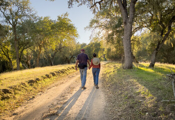 Male and female couple walking together holding hands on romantic stroll on dirt path into the sunset through the oak trees, casting long shadows 