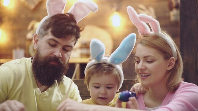 Happy family of mother, father and child son drawing easter eggs at home, painted eggs. Family celebrating easter with kids at home, funny easter rabbits parents with kids in bunny ears.