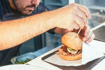 The chef prepares a burger, a hamburger. Delicious and fast food, menu, restaurant and catering, gastronomy. Close up of a male chef, preparing a tasty, fresh, delicious hamburger in a restaurant or p - obrazy, fototapety, plakaty