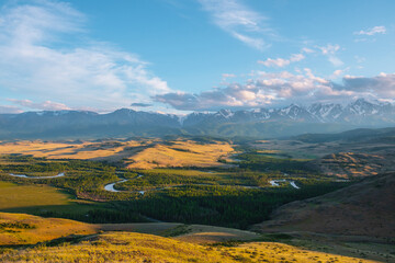 Scenic sunny view from sunlit grassy hill to forest valley with serpentine river against high snowy mountain range in sunlight. Beautiful snake mountain river in forest and snow mountains in sunshine.