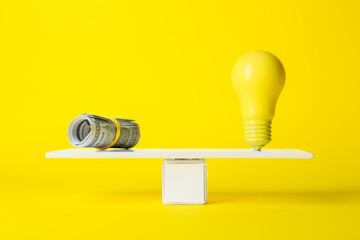 Dollar banknotes and light bulb on teeterboard against yellow background. Concept of balance