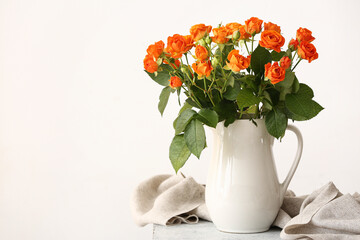 Jug with beautiful orange roses on table against light wall