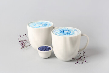 Cups of blue matcha latte, powder and flowers on white background