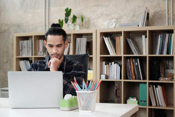 Young male worker of Asian ethnicity uses laptop to do creative work on white desk in front of bookshelf of a casual workplace, startup business person, and online e-commerce occupation. © tigercat_lpg