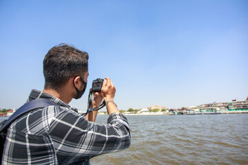 A South Asian tourist, wearing a striped shirt, picked up a camera to photograph Wat Arun on the ferry. and a backpack to walk happily during the long holiday happily in Bangkok, Thailand