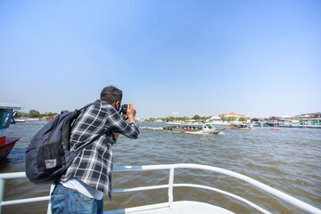 Obraz premium A South Asian tourist, wearing a striped shirt, picked up a camera to photograph Wat Arun on the ferry. and a backpack to walk happily during the long holiday happily in Bangkok, Thailand