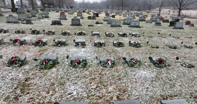 Funeral graveyard cemetery headstone markers decorated with Christmas wreaths during snow storm. Aerial truck shot.
