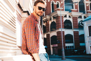 Handsome confident stylish hipster lambersexual model.Modern man dressed in red checkered shirt. Fashion male posing near skyscraper on the street background in sunglasses. Outdoors at sunset