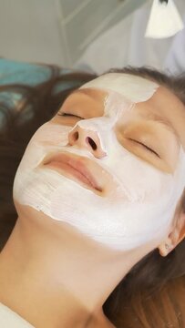 Cosmetologist applies mask to skin of woman's face for therapeutic purposes. Patient is lying on couch and thick white substance is applied to face with brush. Rejuvenation, acne treatment