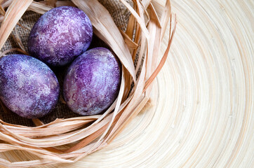 Easter eggs in trendy purple very peri color of the year 2022. Chicken eggs in a straw nest on a wooden plate. Natural dye karkade tea. Zero waste concept. Copy space.	