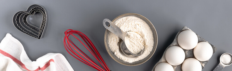 Ingredients for baking on a dark gray background: flour, eggs, baking powder, heart shape, whisk, textiles. top view. Layout of a pie or cake recipe with cookies. Banner