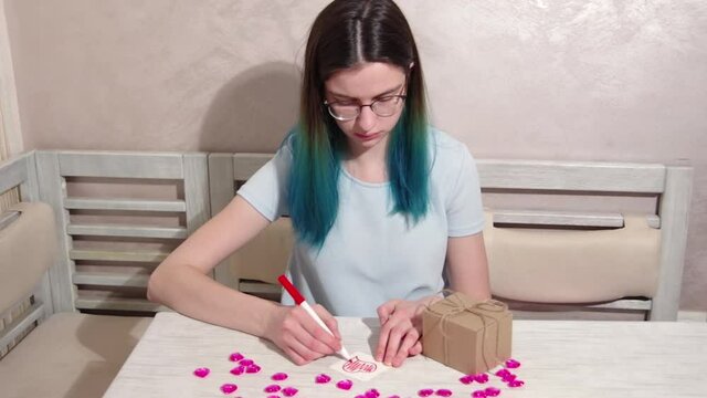 A serious girl with blue hair and glasses decorates a heart shape with a red marker on paper on a table with hearts