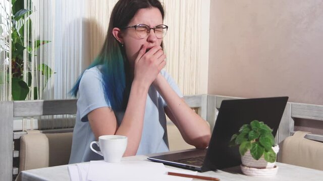 Tired girl student with glasses with blue hair is typing on a laptop, drinking coffee, yawning, full hd.