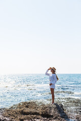 An attractive young woman with long blond hair in a white summer suit stands on the seashore and enjoys the view of the ocean.