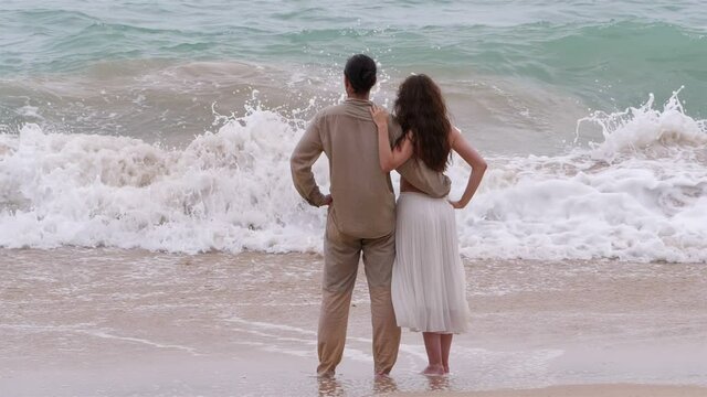 Man and woman look to landswell at sandy beach, telephoto view from back, flatten perspective. Turbulent water surge up and crash down, slow motion shot, couple stand still and stare to sea