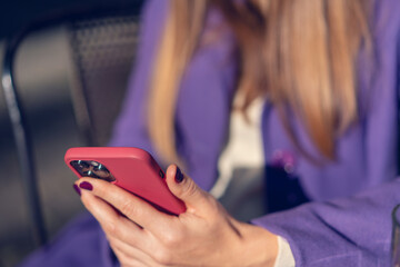A woman in a purple violet coat holding a red smartphone in her hands, close up