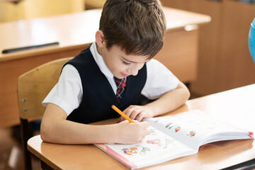 A schoolboy is sitting at a desk in an empty classroom and is writing something in a notebook. School life