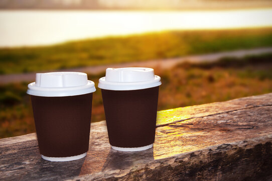 two paper cups with a lid for coffee to take away. Takeaway tea stands on a bench by the riverbank. High quality photo. There is space for text in the background.