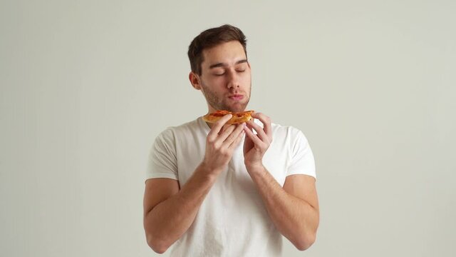 Studio portrait of hungry young man biting into slice of pizza, smelling tainted unappetizing food, dissatisfied with food quality, suffering stomach ache on white isolated background, slow motion.