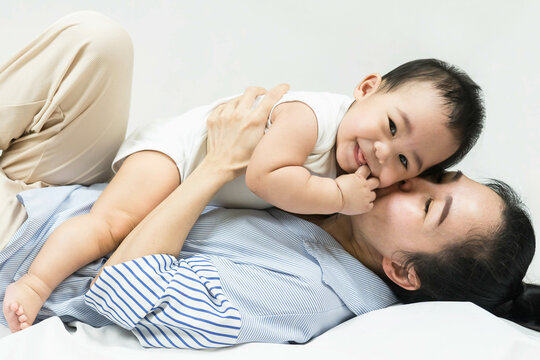 Selective focus Beautiful mom kissing on the cheek newborn with love and caring, Mother showing love to toddler with kiss on cheek.infant baby lying on bed with asian mom.