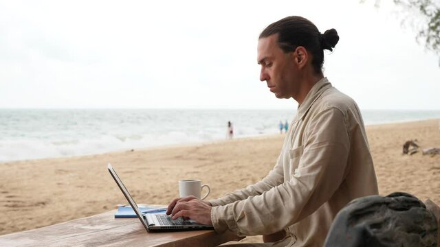 Man work hard, fixed to laptop. He sit at wooden table on sea beach - improvised remote workplace. Tired trope of digital nomad easy lifestyle. Wintering at tropical island of Thailand