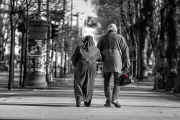 A couple of eldery people in hands are walking through the city street. Rear view.