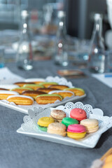Fototapeta na wymiar several colorful macarons in a plate with an openwork napkin. Eclairs with white, brown and green glaze. Gray tablecloth. A plate with eclairs is visible in the background. three glass water bottles