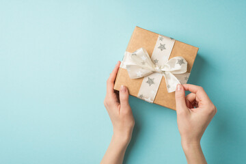 First person top view photo of saint valentine's day decorations young woman's hands untying white ribbon bow with star pattern on kraft paper giftbox on isolated pastel blue background with copyspace
