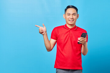 Fototapeta premium Portrait of smiling young asian man using a mobile phone and pointing fingers at copy space isolated on blue background