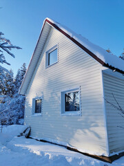 A two-story country house, finished with light siding, on a sunny winter day against a backdrop of snow-covered trees and a blue sky.