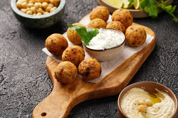 Traditional oriental chickpea deep fried falafel on a wooden board, tzatziki yoghurt sauce, hummus, fresh lime and green cilantro on black surface