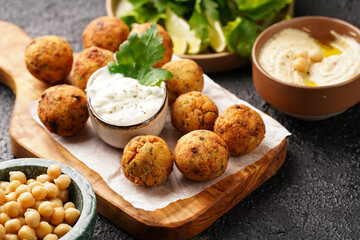 Traditional oriental chickpea deep fried falafel on a wooden board, tzatziki yoghurt sauce, hummus, fresh lime and green cilantro on black surface