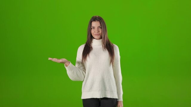 Attractive lady with dark hair tapping twice on empty palm while standing over chroma key background. Young woman in white sweater choosing something with copy space.