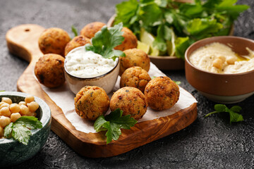Traditional oriental chickpea deep fried falafel on a wooden board, tzatziki yoghurt sauce, hummus, fresh lime and green cilantro on black surface