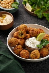 Traditional oriental chickpea deep fried falafel in a deep bowl with tzatziki yoghurt sauce, hummus, fresh lime and green cilantro on black surface