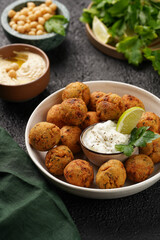 Traditional oriental chickpea deep fried falafel in a deep bowl with tzatziki yoghurt sauce, hummus, fresh lime and green cilantro on black surface
